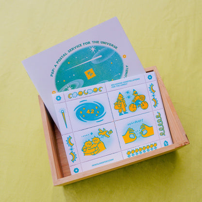 A wooden box containing a zine and a postage stamp sheet of cinderella stamps with pinhole perforation, featuring space-themed illustrations, photographed on a bright yellow-green fabric background.