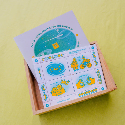 A wooden box containing a zine and a postage stamp sheet of cinderella stamps with pinhole perforation, featuring space-themed illustrations, photographed on a bright yellow-green fabric background.