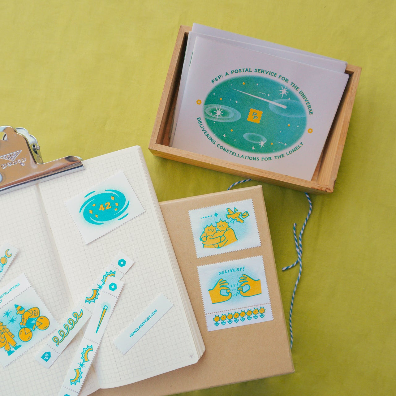 A styled flat-lay of a stationery set on a lime green background, featuring a kraft cardboard box decorated with cinderella stamps.  Decorative stickers, a wooden box containing zines, featuring  a cosmic post office.