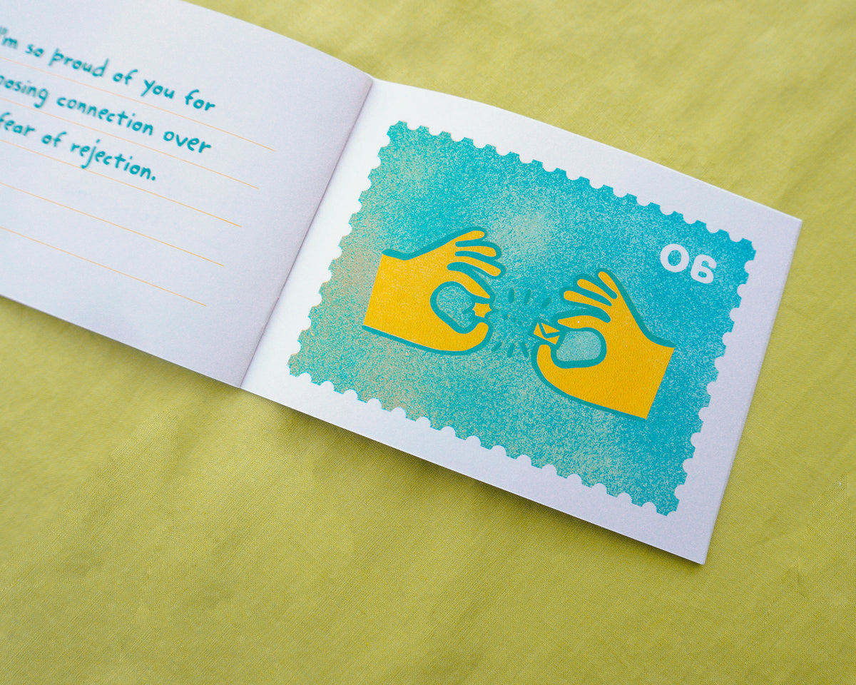 An open zine on a yellow-green textured surface, showing a risograph printed turquoise postage-stamp-style graphic with yellow hand silhouettes and the number 06. The left page has partial handwritten text in blue.