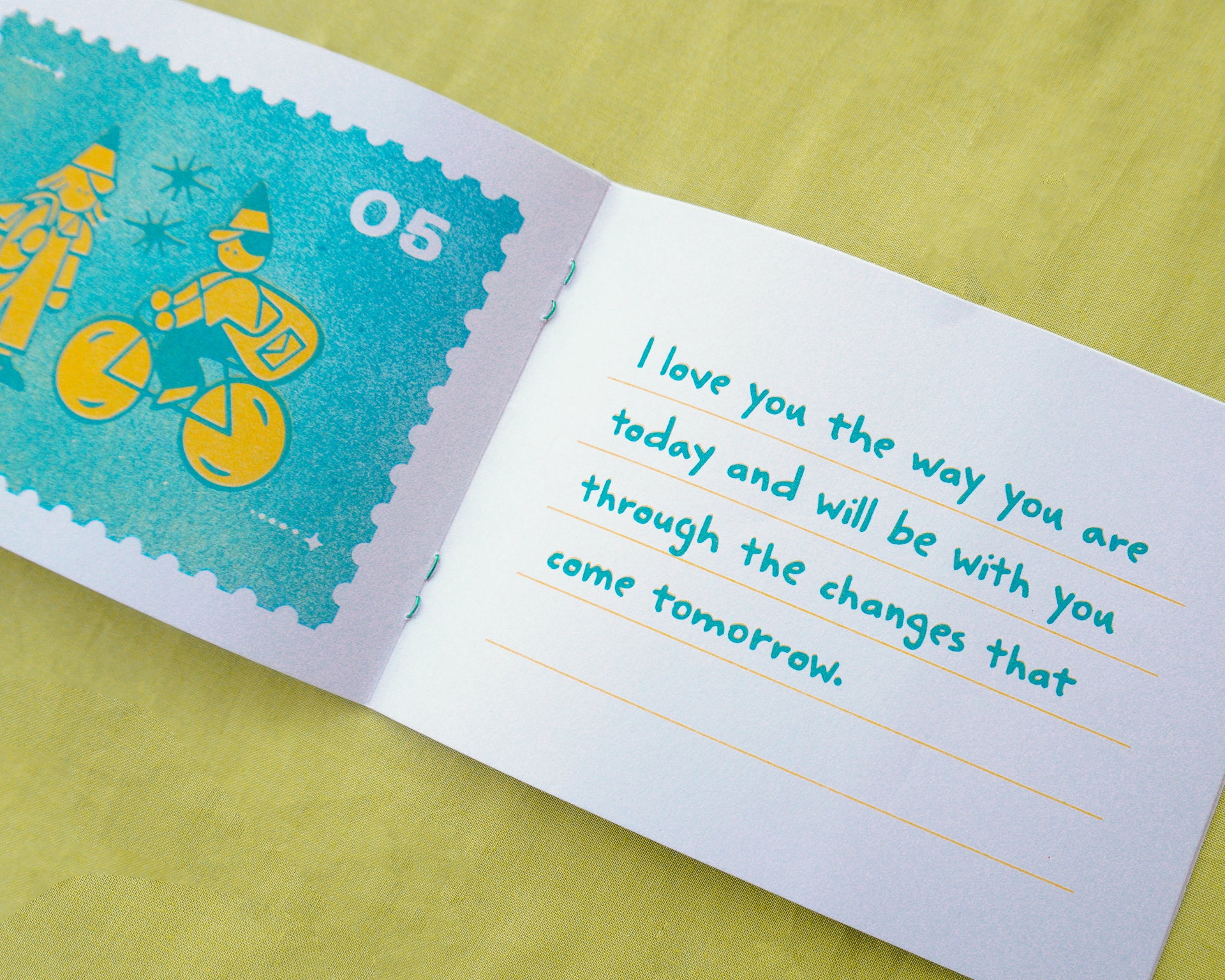 An open white zine or booklet on a yellow-green surface, showing a teal postage-stamp-style illustration on the left page and a handwritten message in teal on the right page.