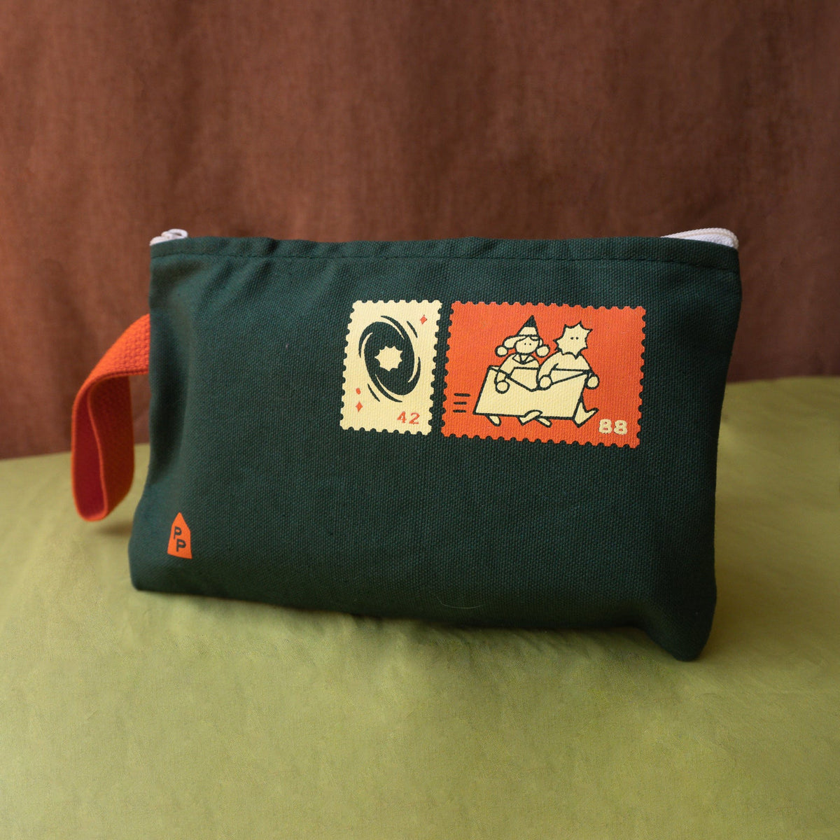 A dark green canvas pouch with a red strap, featuring two vintage-style postage stamp graphics on the front, set against a brown and light green background.