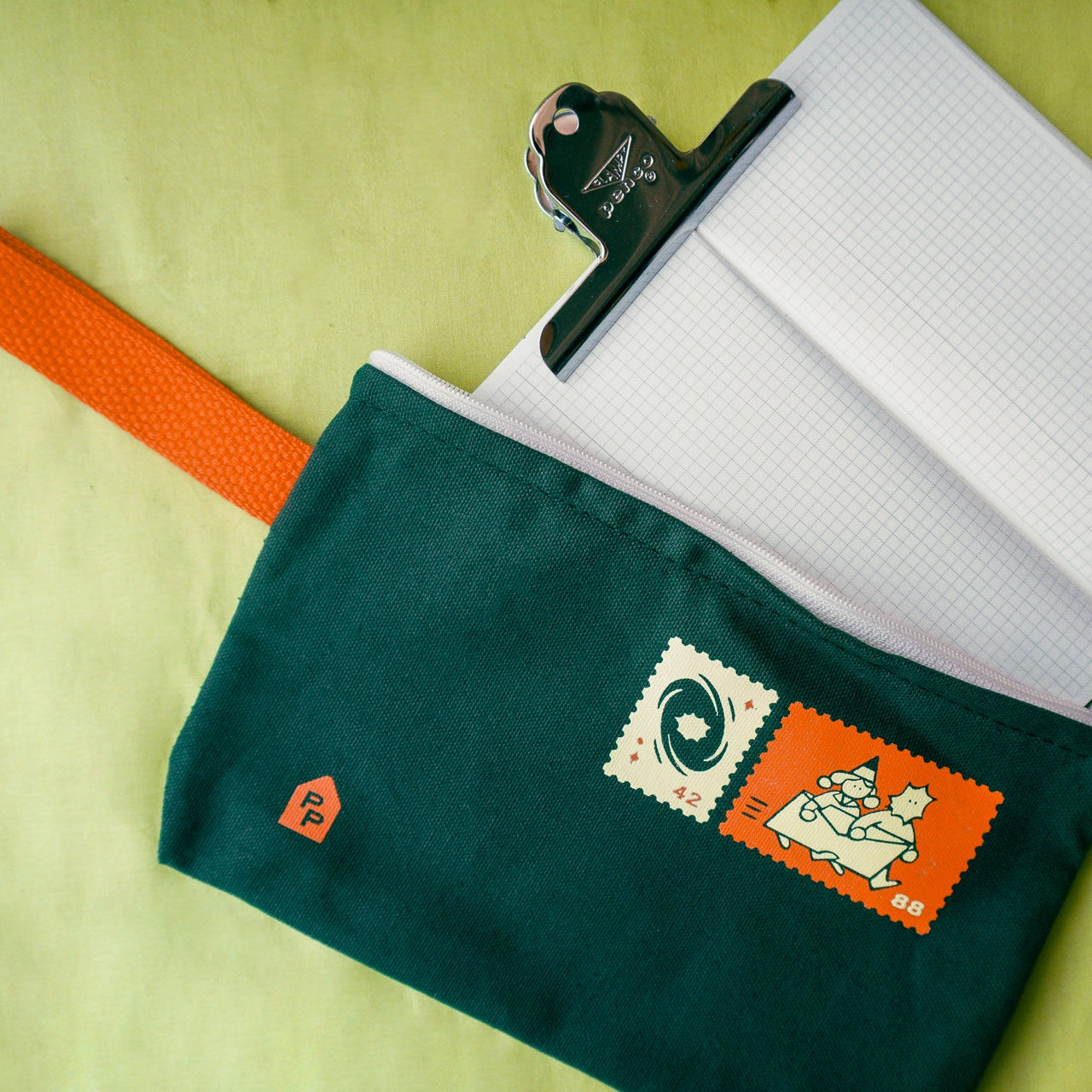A dark green canvas zippered pouch with an orange handle, decorated with two postage stamp graphics, shown with a grid notebook and chrom penco clip on a yellow-green background.