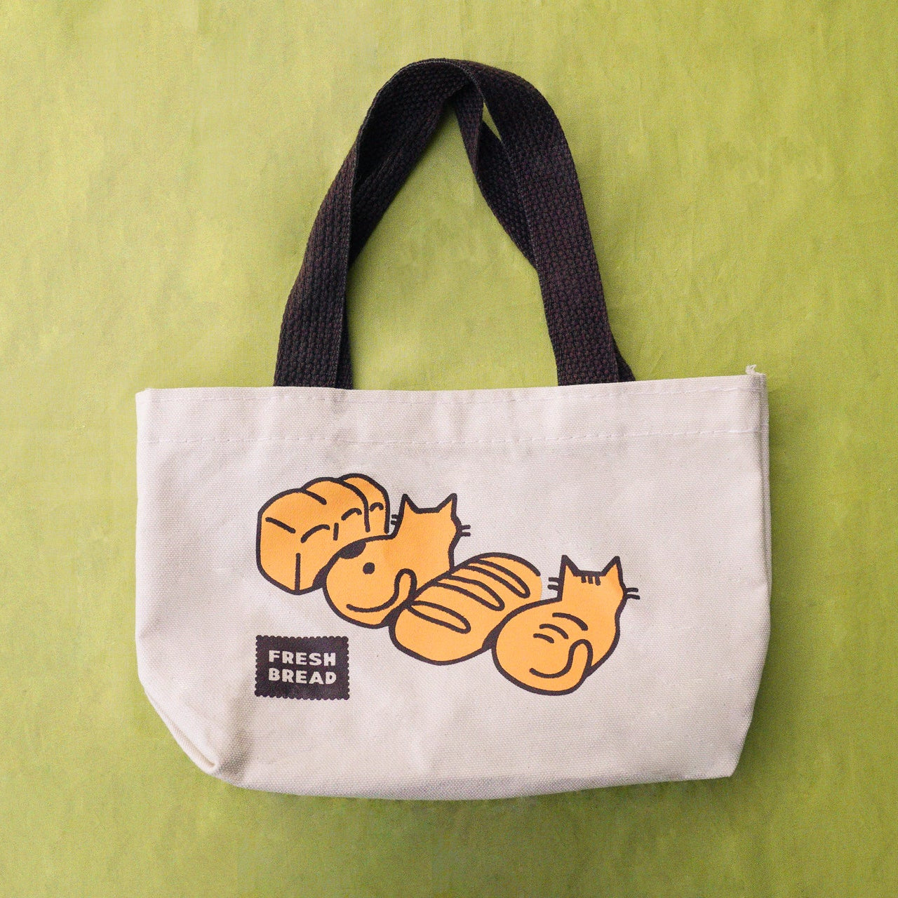 A canvas tote bag with dark brown handles and a graphic of loaves of bread and and cats in loaves, with a small 'FRESH BREAD' label on a postage stamp, set against a textured lime green background.