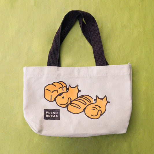 A canvas tote bag with dark brown handles and a graphic of loaves of bread and and cats in loaves, with a small 'FRESH BREAD' label on a postage stamp, set against a textured lime green background.