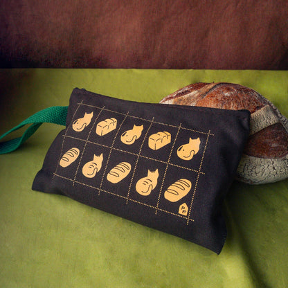 A dark brown fabric pencil pouch with a postage grid of bread illustrations and loafing cats. Featuring a green strap, styled on a green surface with a rustic loaf of bread in the background.