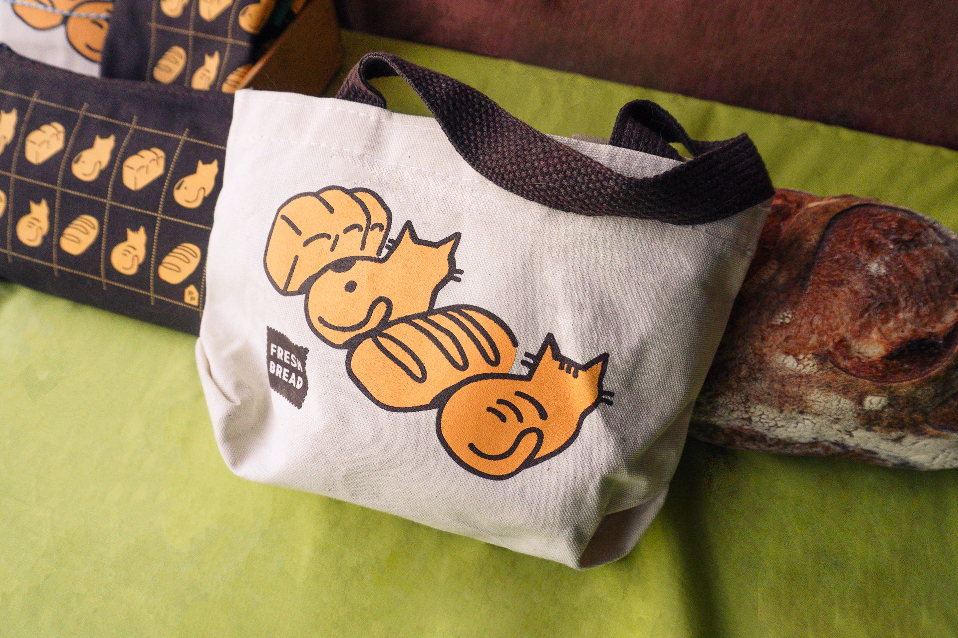 A canvas tote bag with black handles, featuring a graphic of two orange sleeping cats and two loaves of bread. A small 'Fresh Bread' logo, styled on a green surface next to a real rustic loaf of bread.