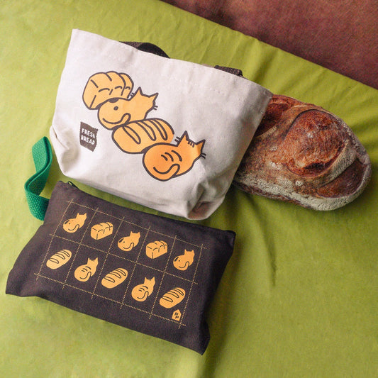 One mini tote and 1 fabric pencil pouch feature cats sitting in loaves and bread illustrations are arranged on a lime green surface next to a real loaf of bread. One pouch is off-white with a green strap, and the other is dark brown.