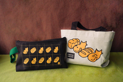 One tote-style bag and one pencil pouch with a wristlet are displayed together on a green surface against a dark brown textured backdrop. The left bag is brown with a yellow bread-grid graphic and a green strap. and the right is on a natural canvas with a brown strap. 