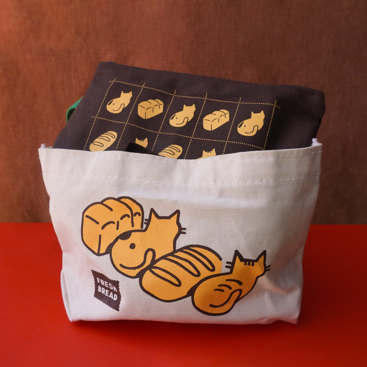 A white canvas tote bag with a cartoon bread and cats illustration sits on a red surface against a brown backdrop, with a dark brown fabric pencil pouch peeking out from the top featuring a grid of similar illustrations.