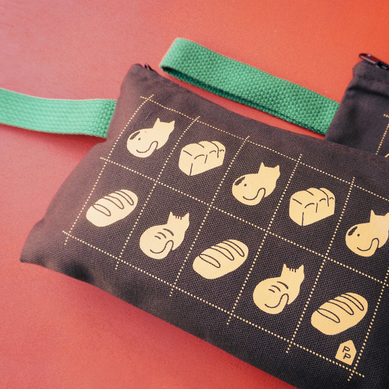 Two brown fabric pouches with green straps, printed with a grid of beige cat and bread illustrations, displayed on a solid red background.