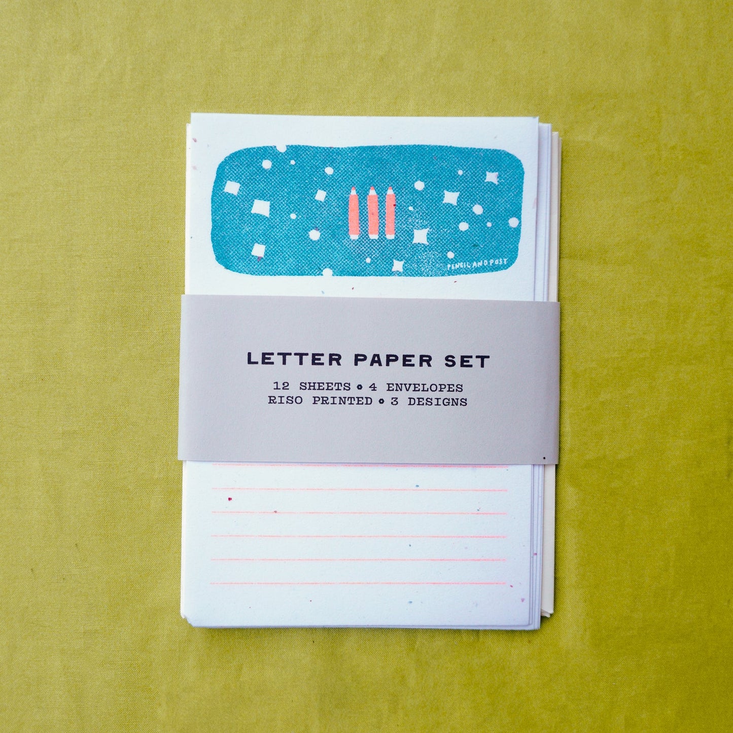 A letter paper set with a gray paper band labeled 'LETTER PAPER SET' wrapped around a stack of white lined sheets and envelopes, placed on a textured yellow-green background.