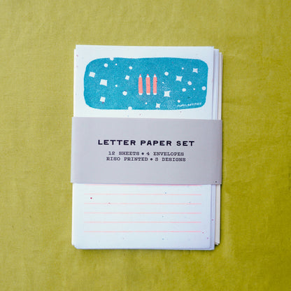 A letter paper set with a gray paper band labeled 'LETTER PAPER SET' wrapped around a stack of white lined sheets and envelopes, placed on a textured yellow-green background.
