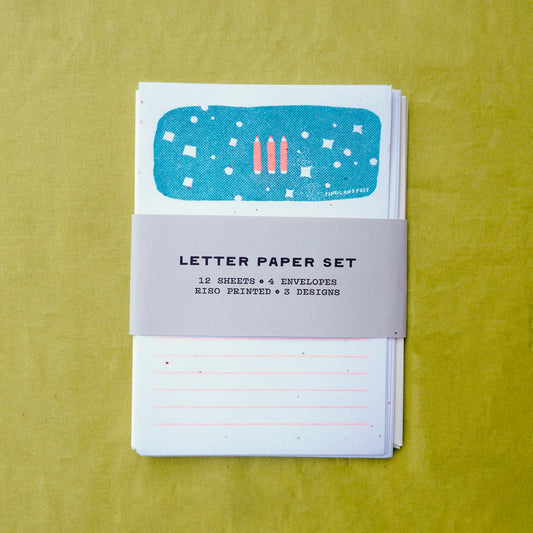 A letter paper set with a gray paper band labeled 'LETTER PAPER SET' wrapped around a stack of white lined sheets and envelopes, placed on a textured yellow-green background.